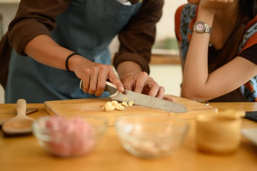 Man in apron chopping garlic on wooden board while preparing meal with his wife in the kitchen.