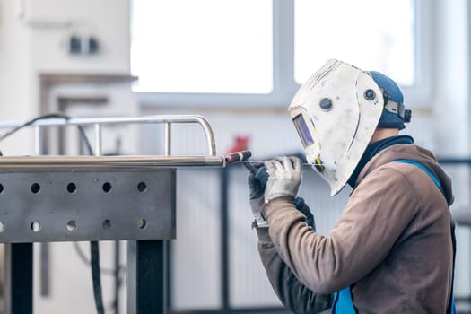 A man in workwear and a welding helmet is using a pneumatic tool to weld a piece of metal, ensuring safety with gloves and gas while building a window in an engineering service