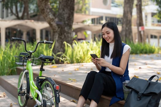 Asian businesswoman with bicycle using smartphone and sitting outside the office building. Woman commuting on bike go to work. Eco friendly vehicle, sustainable lifestyle concept.