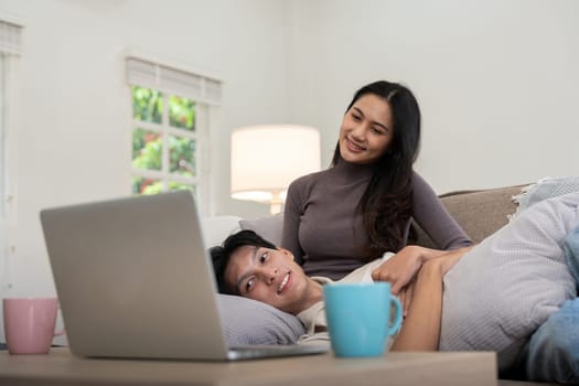 Happy couple asian sit relax on couch in living room watching video on laptop together at home.