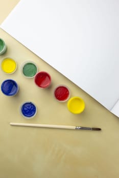 Jars of gouache paint near brush and sketchbook on the table, art concept with vibrant colors and white sheet of paper