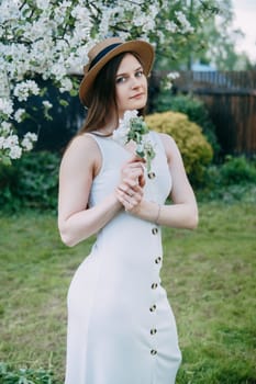 Beautiful young girl in white dress and hat in blooming Apple orchard. Blooming Apple trees with white flowers