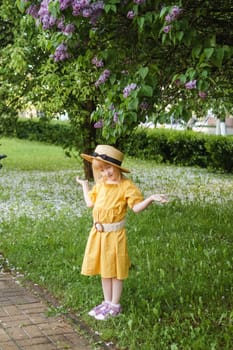 A little girl in a yellow dress and straw hat wearing a bouquet of lilacs. A walk in a spring park, blossoming lilacs