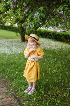 A little girl in a yellow dress and straw hat wearing a bouquet of lilacs. A walk in a spring park, blossoming lilacs