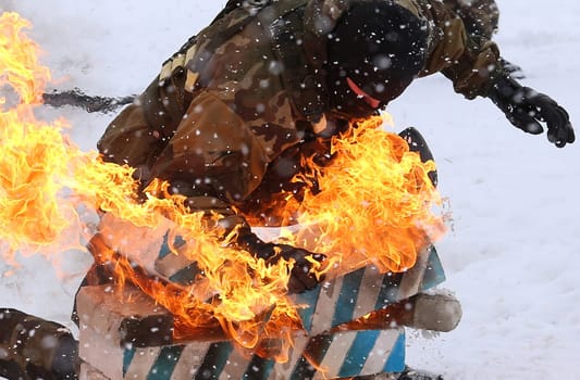 A commando in protective gear fearlessly slides down a snowy slope on a rescue sled, surrounded by flames and smoke, demonstrating courage and skill in this captivating image.