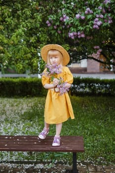 A little girl in a yellow dress and straw hat wearing a bouquet of lilacs. A walk in a spring park, blossoming lilacs