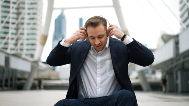 Smart business man wear headphone while sitting at stair in urban city and listen to relaxing music. Happy male leader using headset and moving to song in lively mood surrounded by skyscraper. Urbane.