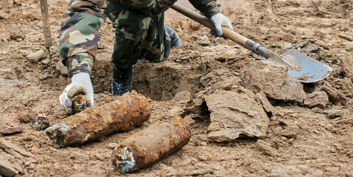 Soldier in protective gear carefully removes live mortar shell from ground during demining operation. Soldier uses shovel to dig around shell. Image shows barren landscape in background.