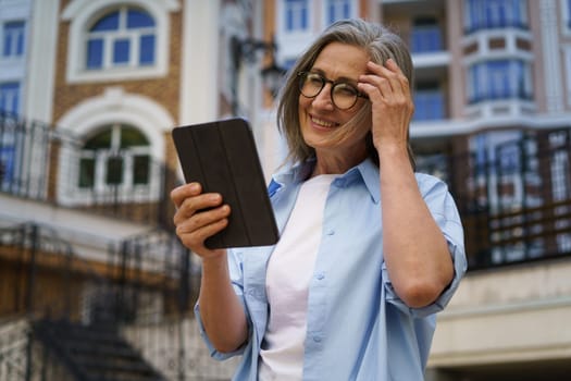 A woman wearing glasses is standing in front of a building, holding a tablet in her hands.