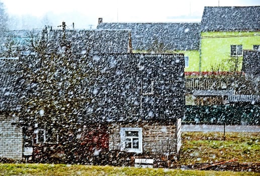 The image shows a small village on a gloomy winter day. The snow is falling heavily, and the houses are covered in snow. The image has a cold and desolate feel to it.