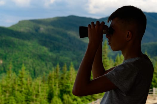Boy teenager looks through binoculars at the mountains, silhouette boy on the background of mountains.