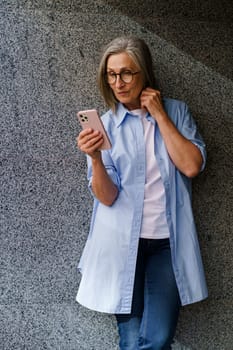 A woman standing against a wall while holding a cell phone in her hand, looking at the screen.