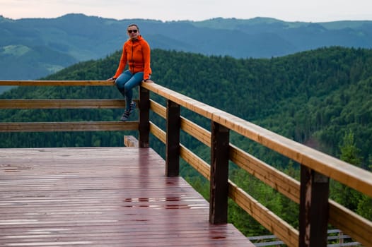 a young girl sits on a wooden handrail, against the background of mountains and evergreen forest.