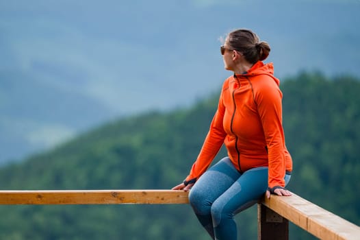 a young girl sits on a wooden handrail, against the background of mountains and evergreen forest.