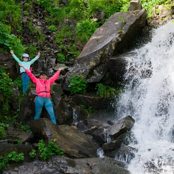 Mother and daughter are standing near a mountain waterfall, active summer vacation.