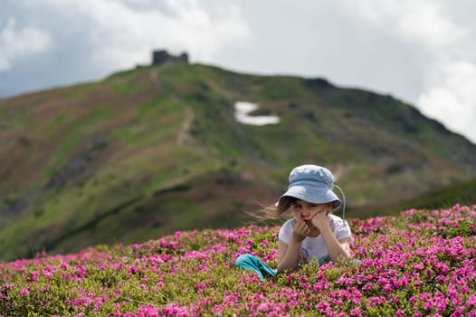 A little girl is sitting on a meadow full of rhododendrons, a beautiful girl among beautiful flowers in the mountains.