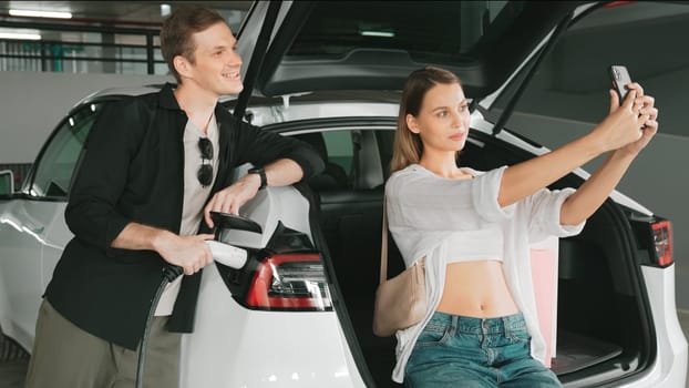 Young couple taking selfie with EV electric car charging battery at shop center parking lot n city downtown as green urban sustainable lifestyle by rechargeable energy of EV vehicle innards