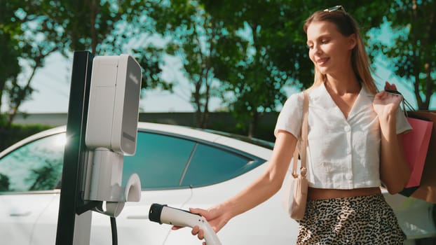 Young woman travel with EV electric car charging in green sustainable city outdoor garden in summer. Urban sustainability lifestyle by green clean rechargeable energy of electric BEV vehicle innards