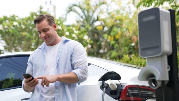 Man use smartphone to pay for electricity at public EV car charging station in nature. Modern environmental and sustainable automobile transportation lifestyle with EV vehicle. Synchronos