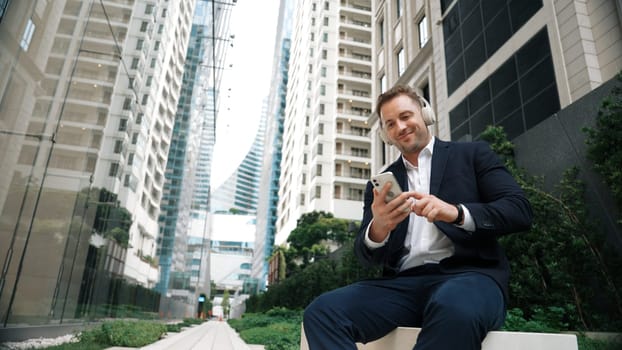 Caucasian businessman listening relaxing music while hold mobile phone. Skilled happy executive manager open music and move to song in lively mood while sitting at green urban city or park. Urbane.