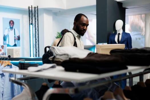 African american man browsing through rack of casual clothes in mall fashion department. Customer choosing outfit and checking apparel on hangers while shopping in boutique