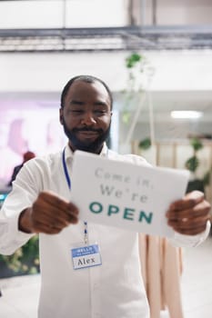 Fashion shop happy african american man assistant hanging opening sign on boutique window. Smiling cheerful clothing store employee holding sign board noticing buyers about work day beginning