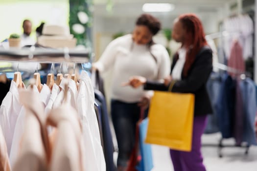 Formal female apparel hanging on metal rack with friends shopping for clothes in mall on blurred background. Trendy shirts and jackets on hangers in fashion boutique close up