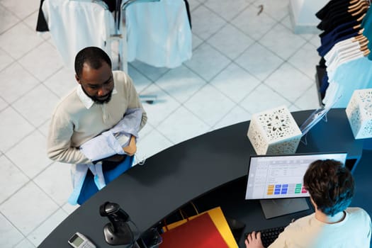 Clothing store worker using cash register software on computer while buyer standing in line at checkout top view. Shopping center employee cashier working at counter desk while customer waiting