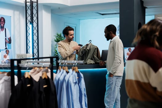 African american man waiting while cashier scanning apparel at clothing store counter desk. Shopping mall boutique client buying trendy garment and standing near cash register