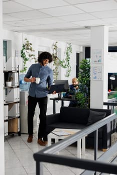 Start up company executive manager working at financial analysis report while standing with laptop near workplace. Arab man drinking coffee and solving business tasks on computer in coworking space