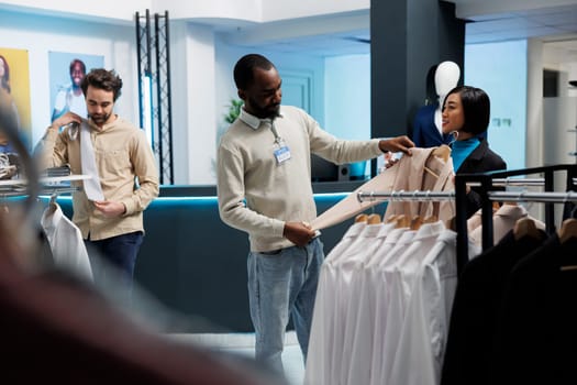 Smiling asian woman holding jacket and asking boutique consultant for fashion advice while shopping for formal outfit. Clothing store client and worker discussing womenswear