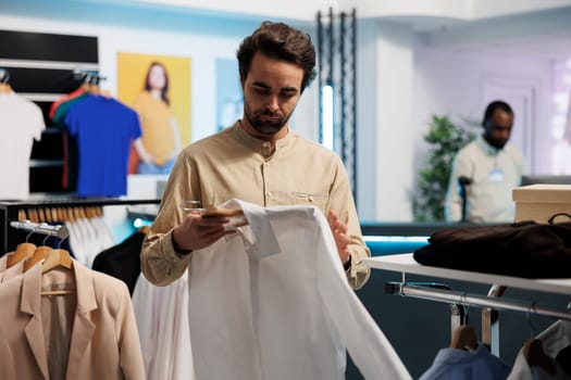 Man checking stylish casual shirt in shopping mall department outlet. Young customer examining trendy garment on hanger while choosing apparel in retail center fashion boutique