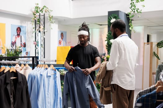 African american buyer checking stylish shirt on hanger in clothing store while assistant giving advice. Customer asking shopping mall employee for help while choosing casual outfit