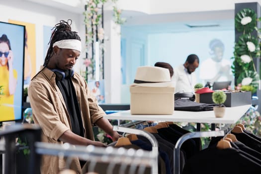 African american man browsing rack with hanging clothes while shopping in fashion boutique. Young buyer choosing stylish outfit from casual menswear new collection in mall