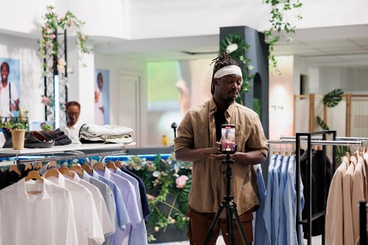 African american influecer promote clothing store while recording video on smartphone. Man creating online social media advertising on mobile phone in shopping mall fashion boutique