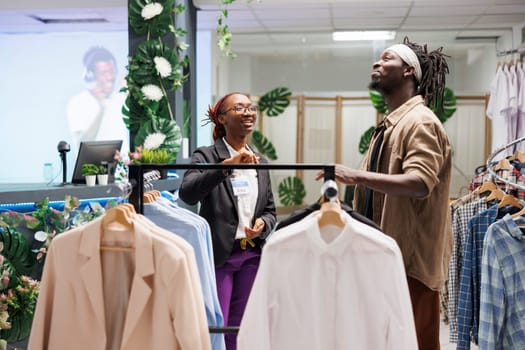 Smiling african american assistant and customer choosing clothes in fashion store. Man customer shopping in mall clothing boutique and asking cheerful woman manager for help