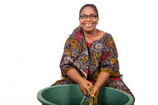 smiling african woman sitting and washing clothes in a big green bowl isolated on white background.