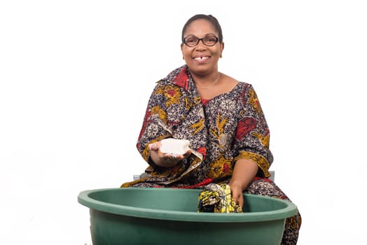 Smiling african woman sitting and washing clothes in a green bowl grade and showing a big piece of soap to the white camera isolated on white background.