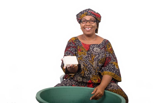 Smiling african woman sitting and washing clothes in a green bowl grade and showing a big piece of soap to the white camera isolated on white background.