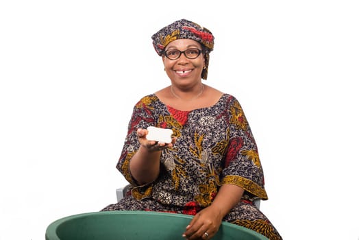 Smiling african woman sitting and washing clothes in a green bowl grade and showing a big piece of soap to the white camera isolated on white background.