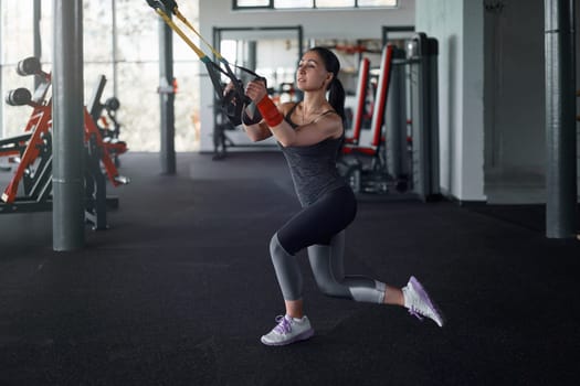 Full body of sportswoman exercising training with suspension straps. Young female athlete pulling special resistance straps in gym. Fitness and health concept.