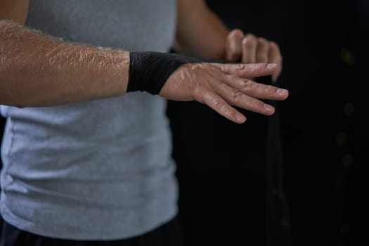 Crop unrecognizable male kickboxer wrapping bandage before training. Sportsman wearing black tape on hand against dark background.