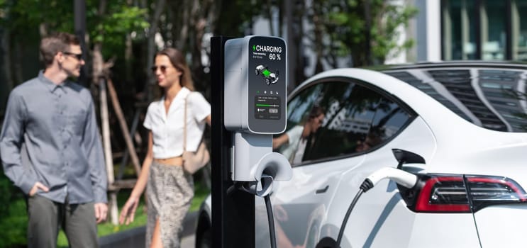 Young couple travel with EV electric car charging in green sustainable city outdoor garden in summer shows urban sustainability lifestyle by green clean rechargeable energy of electric vehicle innards