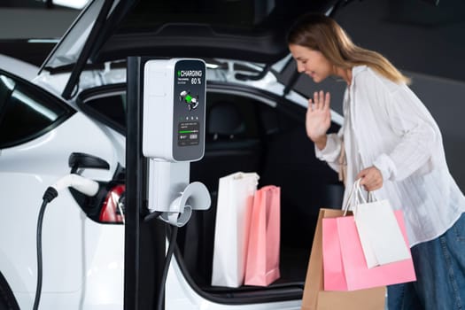 Young woman travel with EV electric car to shopping center parking lot charging in downtown city showing urban sustainability lifestyle by green clean rechargeable energy of electric vehicle innards