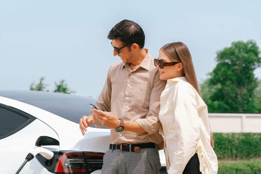 Young couple use smartphone to pay for electricity at public EV car charging station green city park. Modern environmental and sustainable urban lifestyle with EV vehicle. Expedient