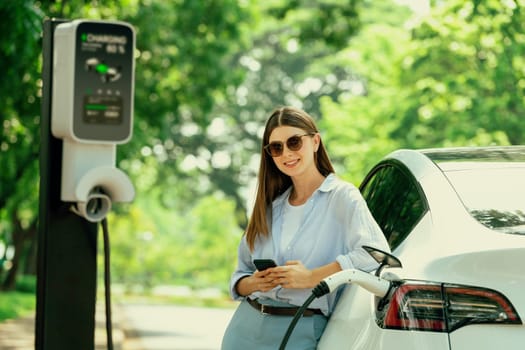 Young woman using smartphone online banking application to pay for electric car battery charging from EV charging station during vacation holiday road trip at national park or summer forest. Exalt