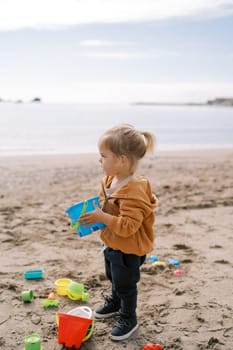 Little girl with a toy bucket in her hands stands on a sandy beach surrounded by colorful toys. High quality photo
