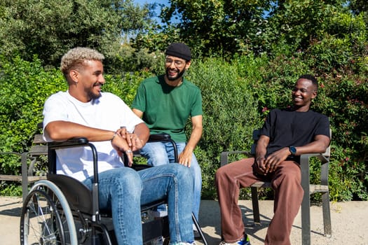 Young disabled African American man in a wheelchair and his male friends talking and having fun together sitting on a bench in city park. Friendship concept.