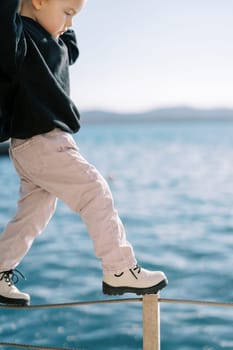 Little girl walks along a rope fence by the sea, looking at her feet. High quality photo