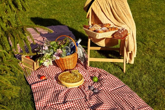 Cozy summer picnic scene on green grass with homemade quiche, bottle of wine in wicker basket and fresh fruits scattered on checkered blanket, fluffy croissants and chocolate roll cake on wooden chair